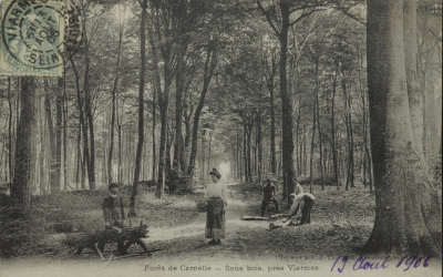 Carte postale en noir et blanc montrant une forêt, dans laquelle travaillent des bûcherons. Une femme se tient debout avec un panier à la main.